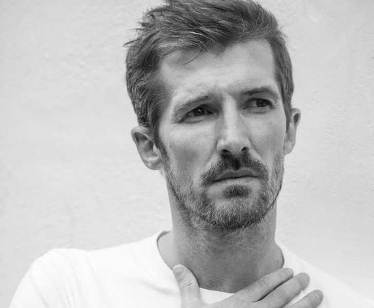 Picture of Gwilym Lee posing for a photo by putting his right hand on his neck in black and white themed picture with white wall on background